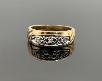 May include: A gold ring with a rectangular face featuring three small, clear stones set in a decorative pattern. The band has a ribbed design. The ring is displayed on a reflective surface.