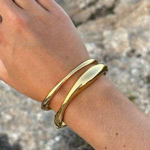 May include: A hand adorned with a gold ring featuring a pale blue gemstone and two gold bangles. The bracelets have a smooth, polished surface. The jewellery is set against a natural, outdoor backdrop.
