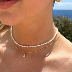 May include: A gold chain necklace with a small letter 'A' pendant and a pearl choker necklace. The necklace is worn by a person with a gold earring.