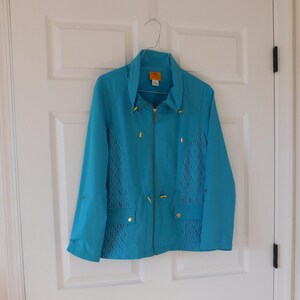 May include: A turquoise jacket with a stand-up collar and a zipper closure. The jacket features gold-colored accents on the zipper, pockets, and drawstrings. The front panels have a textured design. The jacket is hanging on a metal hanger.