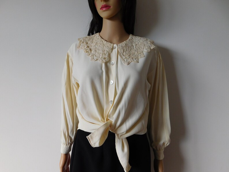Puede incluir: Una blusa de color crema con cuello de encaje y mangas largas. La blusa est&aacute; anudada a la cintura y tiene un cierre de botones en la parte delantera.