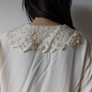 Puede incluir: Una blusa blanca con un delicado cuello de encaje. El cuello es un dise&ntilde;o grande e intrincado con un borde festoneado.