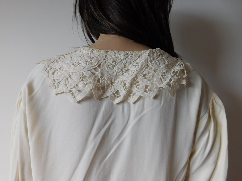 Puede incluir: Una blusa blanca con un delicado cuello de encaje. El cuello es un dise&ntilde;o grande e intrincado con un borde festoneado.