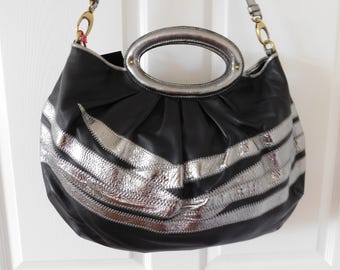 May include: A black hobo bag featuring a metallic silver oval handle and horizontal silver textured stripes across the front. The bag includes a detachable shoulder strap with gold-toned hardware clips and a gathered, pleated design.
