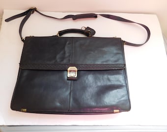 May include: A dark brown leather briefcase featuring a top handle, an adjustable shoulder strap, and a front flap with a metallic push-lock closure. The flap has a textured border, and the bag includes small metallic corner accents.