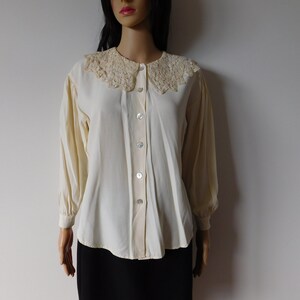 Puede incluir: Una blusa blanca de botones con un delicado cuello de encaje. La blusa tiene mangas largas y un corte holgado.