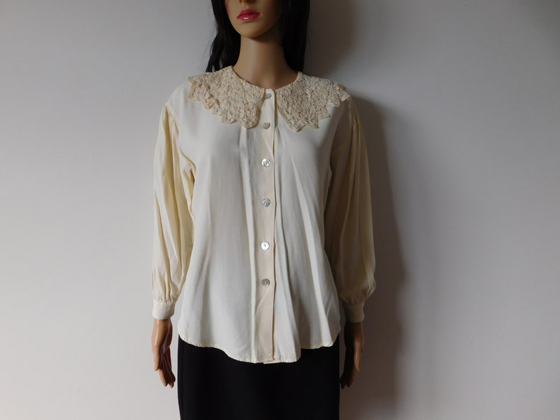 Puede incluir: Una blusa blanca de botones con un delicado cuello de encaje. La blusa tiene mangas largas y un corte holgado.