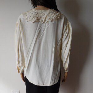 Puede incluir: Una blusa blanca de manga larga con un delicado cuello de encaje. La blusa tiene un corte holgado y un dobladillo ligeramente redondeado.