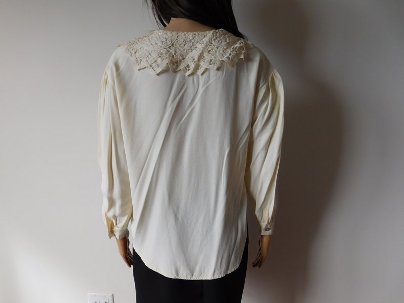 Puede incluir: Una blusa blanca de manga larga con un delicado cuello de encaje. La blusa tiene un corte holgado y un dobladillo ligeramente redondeado.