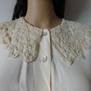 Puede incluir: Una blusa blanca con un gran cuello de encaje de color crema. El cuello tiene un borde festoneado y est&aacute; hecho de encaje delicado e intrincado.