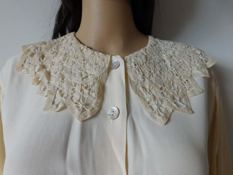 Puede incluir: Una blusa blanca con un gran cuello de encaje de color crema. El cuello tiene un borde festoneado y est&aacute; hecho de encaje delicado e intrincado.