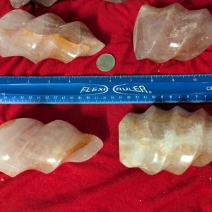 May include: Four natural quartz crystals with a swirling, layered pattern in shades of white and orange. The crystals are shaped like a spiral and are arranged on a red surface. A ruler is included for scale.