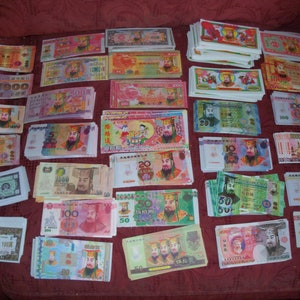 May include: A collection of colorful joss paper banknotes, also known as spirit money, featuring various denominations and portraits of historical figures. The rectangular paper bills are arranged in multiple stacks, displaying vibrant hues of pink, green, blue, and yellow with intricate traditional Chinese text and decorative borders.