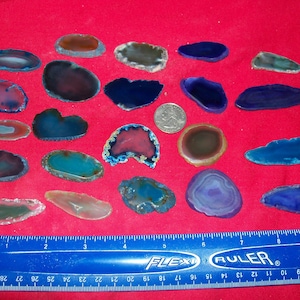 May include: A collection of 20 agate slices in various shades of blue, purple, green, and brown. The slices are arranged on a red surface with a ruler for scale.
