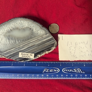 May include: A gray and white agate slice with black banding. The slice is 5.5 inches long, 3.5 inches wide, and 1 9/16 inches thick. It weighs 1 pound and is made in Brazil.