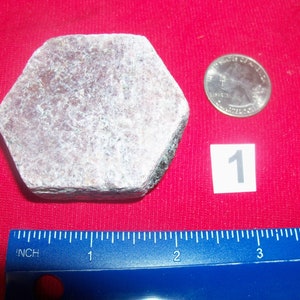 May include: A hexagonal, light purple gemstone with a rough, textured surface. The gemstone is about 1 inch in diameter and is next to a US quarter for size comparison. The number "1" is written on a white piece of paper next to the coin.