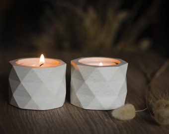May include: Two white geometric concrete candle holders with lit tea lights on a wooden surface.