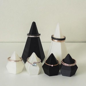 Peut inclure: Ensemble de six présentoirs à anneaux en forme de pyramides, noirs et blancs. Les présentoirs sont disposés en rangée sur une surface blanche. Chaque présentoir est surmonté d'une bague.