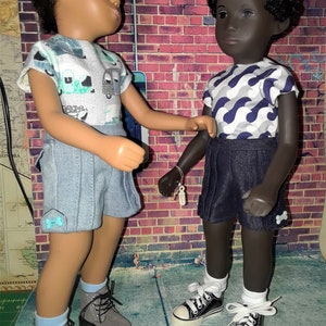 May include: Two dolls, one with brown hair and blue eyes wearing a light blue and white patterned shirt and denim shorts, and the other with black hair and brown eyes wearing a grey and blue patterned shirt and dark blue shorts. Both dolls are wearing white socks and shoes.