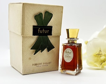 May include: A vintage perfume bottle and box set. The perfume bottle is a small, square shape with a gold cap. The box is beige with a green ribbon design and the word "futur" on it. The box also has the words "Robert Piguet Paris - France".