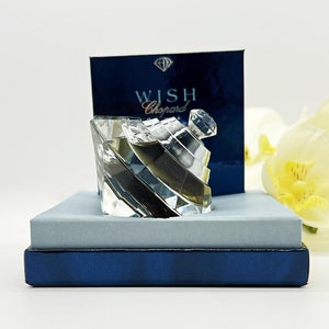 Könnte beinhalten: Ein klares Glasfläschchen Chopard Wish Parfum in seiner blauen Box. Das Fläschchen ist diamantförmig und hat einen silbernen Verschluss. Auf der Box steht "Wish Chopard" gedruckt.
