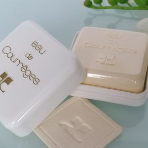Puede incluir: Una caja blanca con un logotipo dorado y el texto "eau de Courr&egrave;ges" en la parte superior. La caja est&aacute; abierta y una pastilla de jab&oacute;n blanca con el texto "eau de Courr&egrave;ges" est&aacute; dentro.