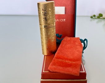 May include: A gold-colored perfume bottle with a floral design and the word "Lalique" engraved on it. The bottle is in a red box with a matching red velvet pouch.