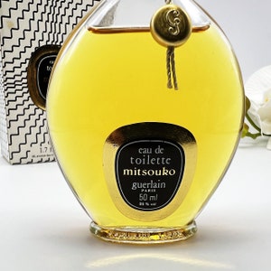 May include: A clear glass bottle of Mitsouko Eau de Toilette by Guerlain. The bottle is shaped like a heart and has a gold cap with a G on it. The label is gold with black text that reads "eau de toilette mitsouko guerlain paris 50 ml 86% vol".