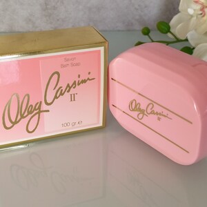 May include: A pink bar of soap in a plastic container with gold lettering that reads "Oleg Cassini II" and "Savon Bain Soap 100 gr. e".