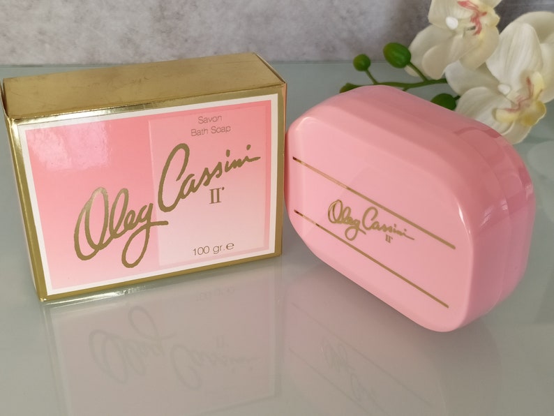 May include: A pink bar of soap in a plastic container with gold lettering that reads "Oleg Cassini II" and "Savon Bain Soap 100 gr. e".