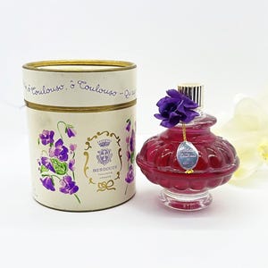May include: A vintage perfume bottle with a deep red hue, topped with a purple flower and a silver cap. The bottle is next to a cylindrical box with floral illustrations and the text "BERDOUES TOULOUSE FRANCE".