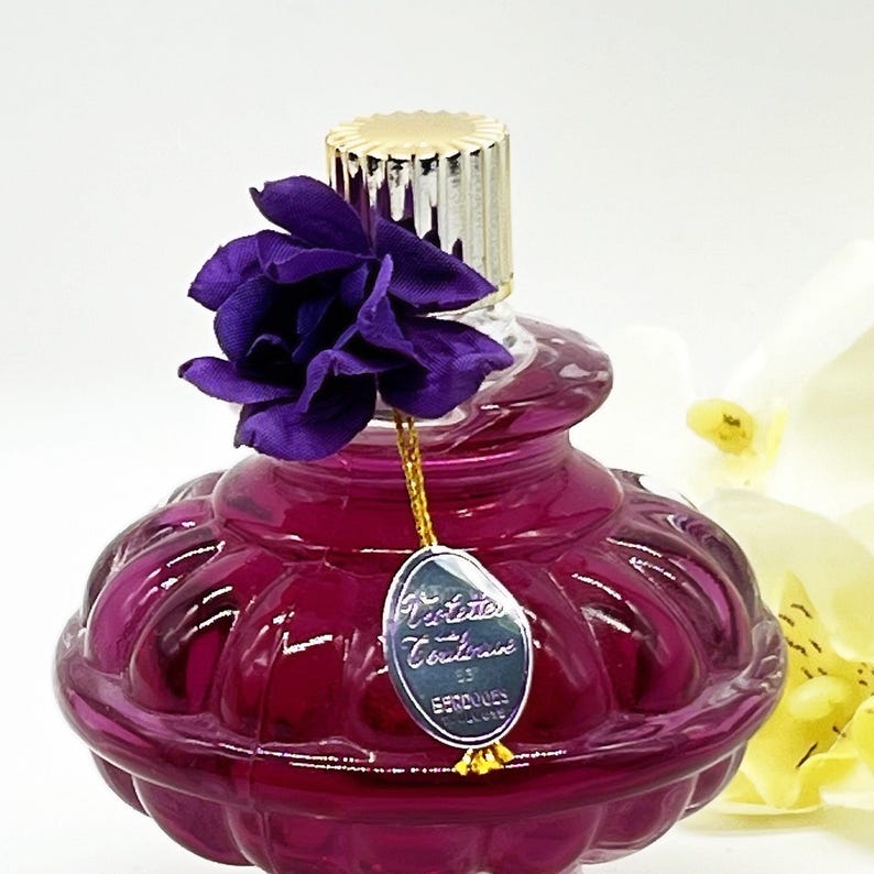 May include: A decorative perfume bottle with a deep purple hue, featuring a gold-colored cap and a purple fabric flower. A small, silver tag hangs from the bottle. The bottle has a unique, rounded shape.