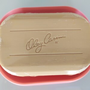May include: A pink soap dish containing a rectangular bar of soap. The soap is wrapped in plastic and has the words "Oleg Cassini" printed on it.