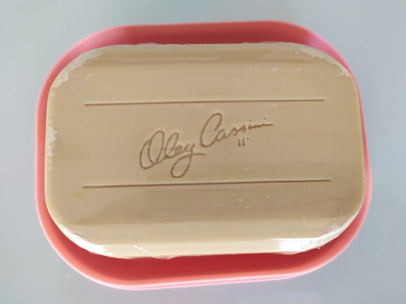 May include: A pink soap dish containing a rectangular bar of soap. The soap is wrapped in plastic and has the words "Oleg Cassini" printed on it.