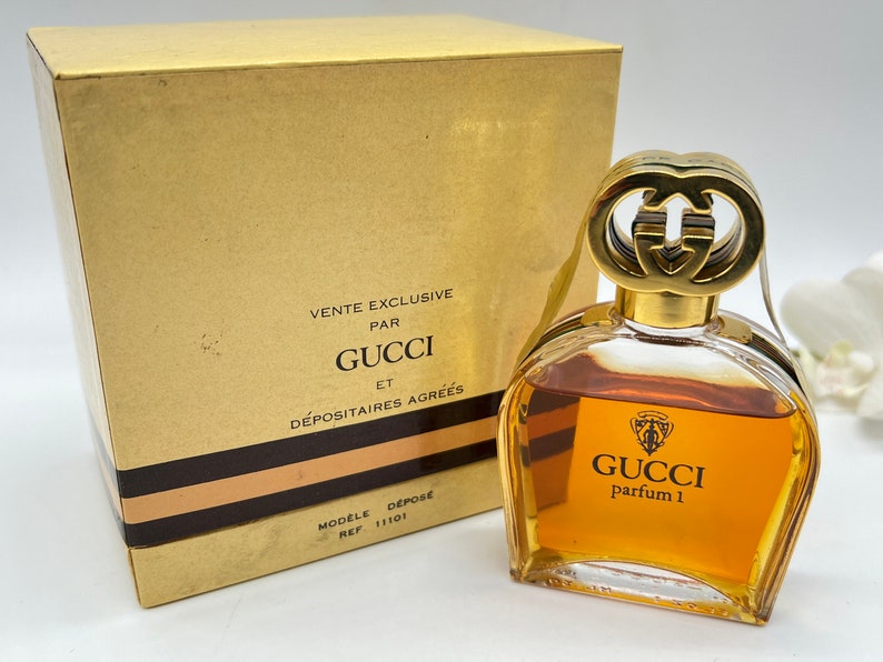 May include: A gold and brown box with the text "Vente Exclusive Par Gucci Et D&eacute;positaire Agr&eacute;es" and a clear glass bottle of Gucci perfume with a gold cap and the text "Gucci Parfum 1".