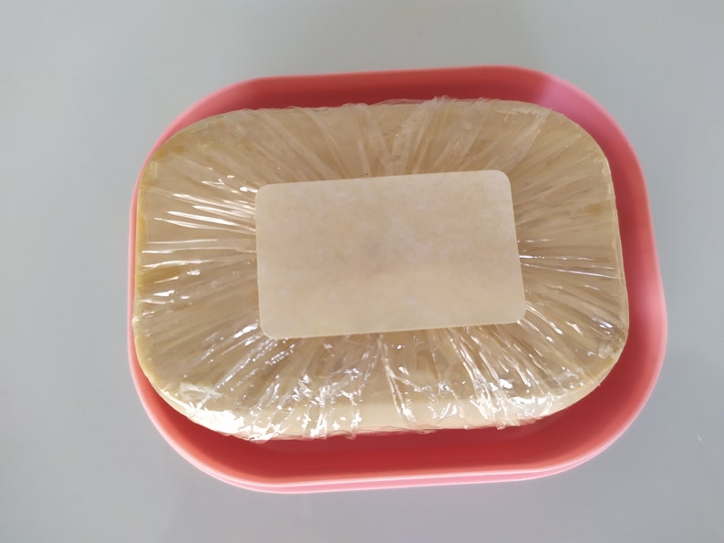 May include: A bar of soap wrapped in plastic and a label, sitting in a pink soap dish.