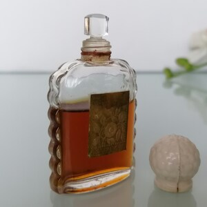May include: A vintage glass bottle of perfume with a clear glass stopper and a gold label. The bottle has a textured design and is filled with a brown liquid. A white ceramic stopper is also pictured.