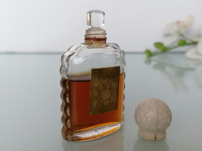 May include: A vintage glass bottle of perfume with a clear glass stopper and a gold label. The bottle has a textured design and is filled with a brown liquid. A white ceramic stopper is also pictured.