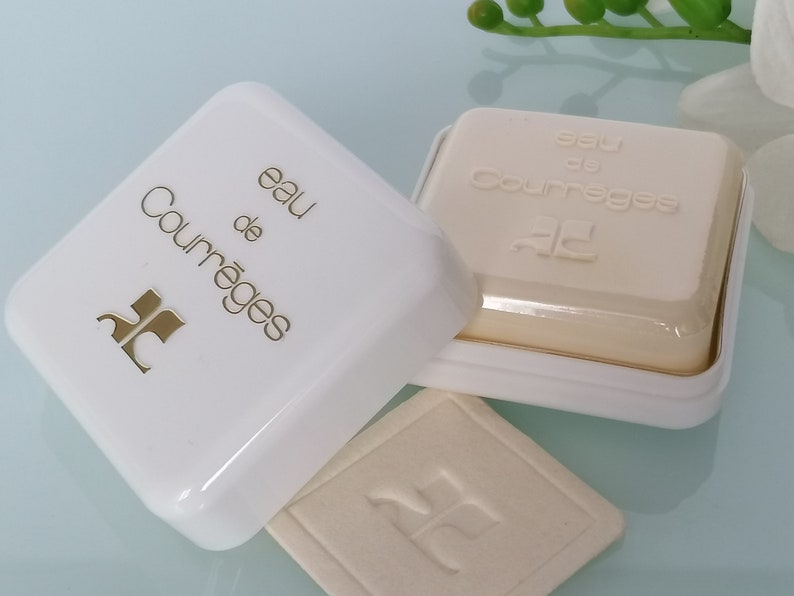 Puede incluir: Una caja blanca con un logotipo dorado y el texto "eau de Courr&egrave;ges" en la parte superior. La caja est&aacute; abierta y una pastilla de jab&oacute;n blanca con el texto "eau de Courr&egrave;ges" est&aacute; dentro.