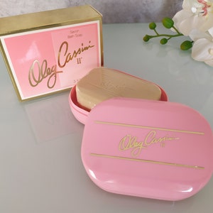 May include: A pink soap dish with a gold inscription that reads "Oleg Cassini II" and a bar of soap inside. The soap dish is on a white surface with a white orchid in the background.
