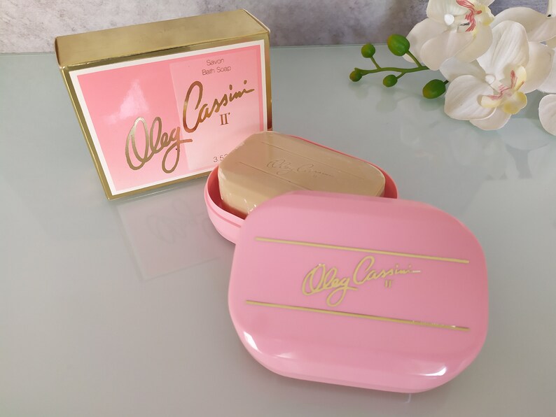May include: A pink soap dish with a gold inscription that reads "Oleg Cassini II" and a bar of soap inside. The soap dish is on a white surface with a white orchid in the background.