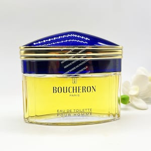 May include: A clear glass bottle of Boucheron Paris Eau de Toilette Pour Homme. The bottle has a gold cap with a blue top and a gold band around the middle. The bottle is sitting on a white surface.