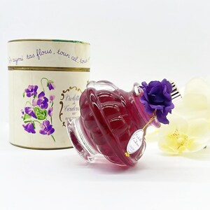 May include: A clear glass perfume bottle filled with a deep red liquid, adorned with a purple flower and a gold tag. The bottle is next to a cylindrical box with floral illustrations and French text. The overall theme is vintage fragrance.