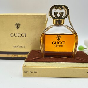 May include: A gold and brown perfume bottle with a gold double G logo on the stopper. The bottle is labeled "GUCCI parfum 1" and is sitting on a brown velvet box. The box is labeled "NET. FL. OZ. 1" and "ML. 30".