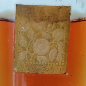 May include: A small, clear glass bottle with a gold label. The label has a floral design with a white rose in the center. The bottle is filled with a dark amber liquid.
