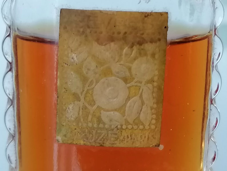 May include: A small, clear glass bottle with a gold label. The label has a floral design with a white rose in the center. The bottle is filled with a dark amber liquid.
