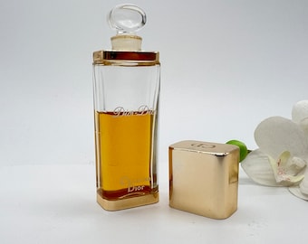 May include: A vintage glass bottle of Dior-Dior perfume with a gold cap and a gold label. The bottle is filled with a golden liquid. The cap is separate from the bottle and is sitting on a white surface.