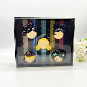 May include: A set of six colourful, round, plastic figurines of women with different hairstyles and facial features. The figurines are arranged in a clear plastic box with a black background. The box has a label with the text "JUKUS LOVERS HARAJUKU" printed on it.