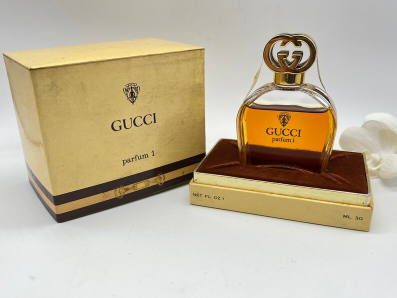 May include: A gold and brown perfume bottle with a gold double G logo on the stopper. The bottle is labeled "GUCCI parfum 1" and is sitting on a brown velvet cushion inside a gold box. The box is labeled "NET FL OZ 1" and "ML 30".