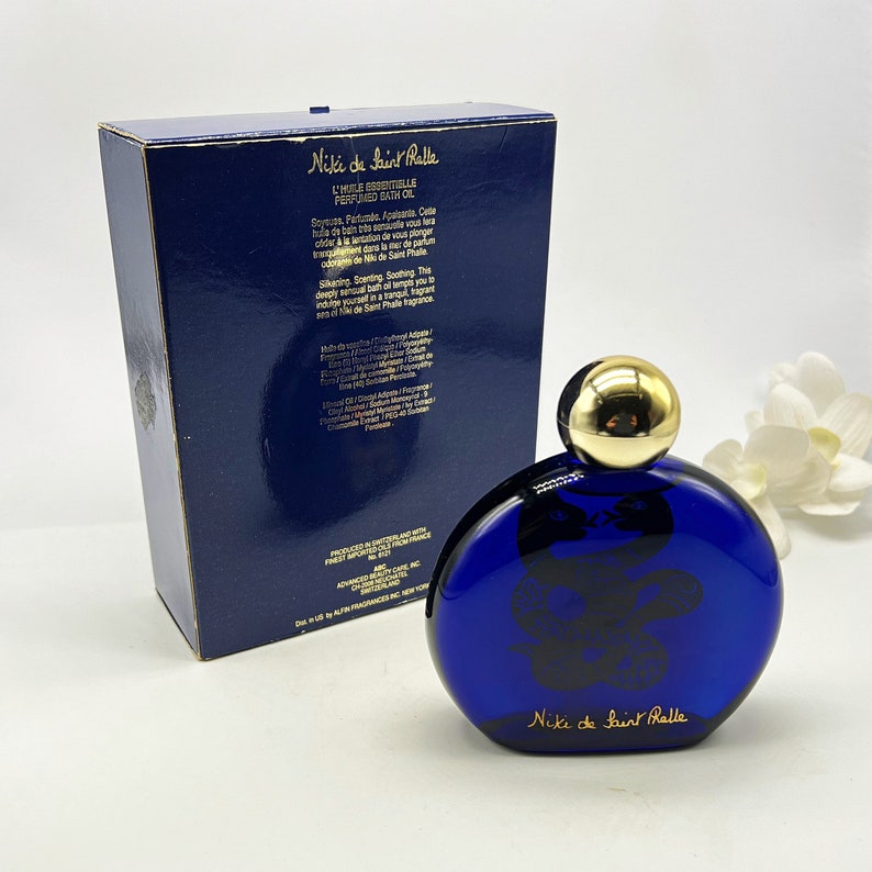 Puede incluir: Una botella de vidrio azul de aceite de ba&ntilde;o perfumado Niki de Saint Phalle. La botella tiene una tapa dorada y un dise&ntilde;o de serpiente en la parte delantera. La botella est&aacute; sobre una superficie blanca.